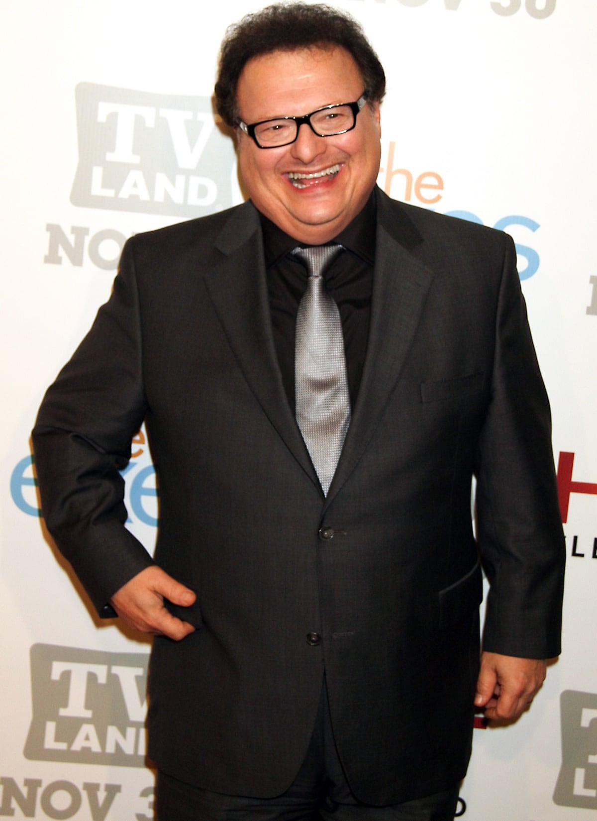 Wayne Knight flashes a big smile in a sleek charcoal suit paired with a silver tie, exuding confidence and style
