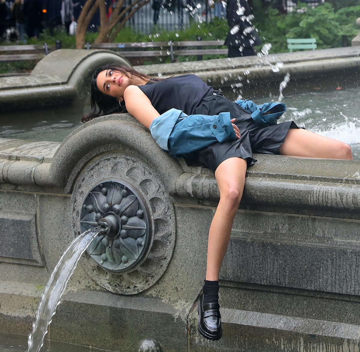 Kendall Jenner leans on the edge of a public fountain in wet clothes that included a pair of black shorts, a tank top, and a pale blue coat