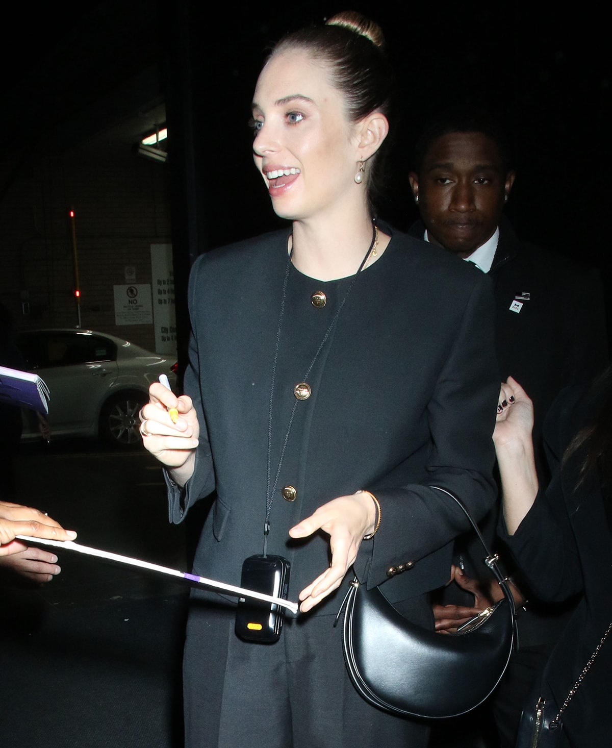 Maya Hawke styles her black ensemble with a black Prada shoulder bag, black Prada pumps, and mismatched pearl earrings