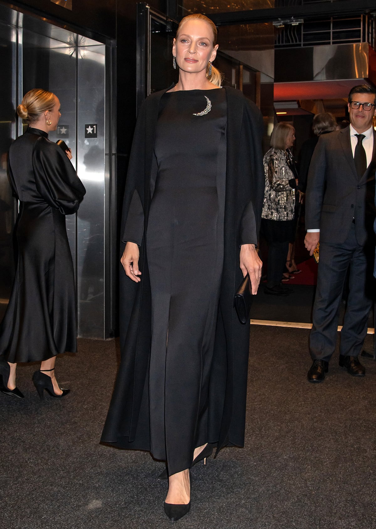 Uma Thurman looks regal in a black column dress with a moon crescent brooch, a matching cape, and black pumps