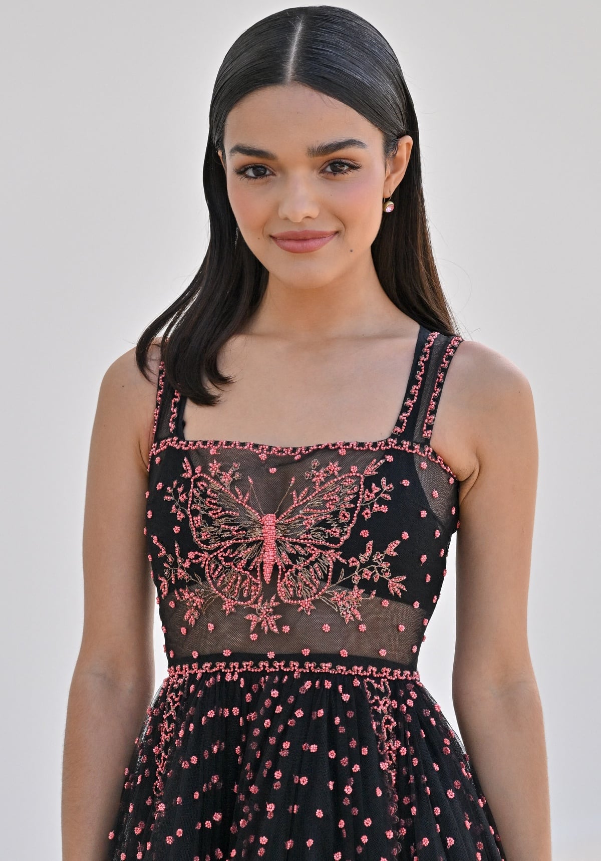 Rachel Zegler wearing a black and pink butterfly-embellished gown with minimal jewelry, sleek black hair, and a simple makeup palette