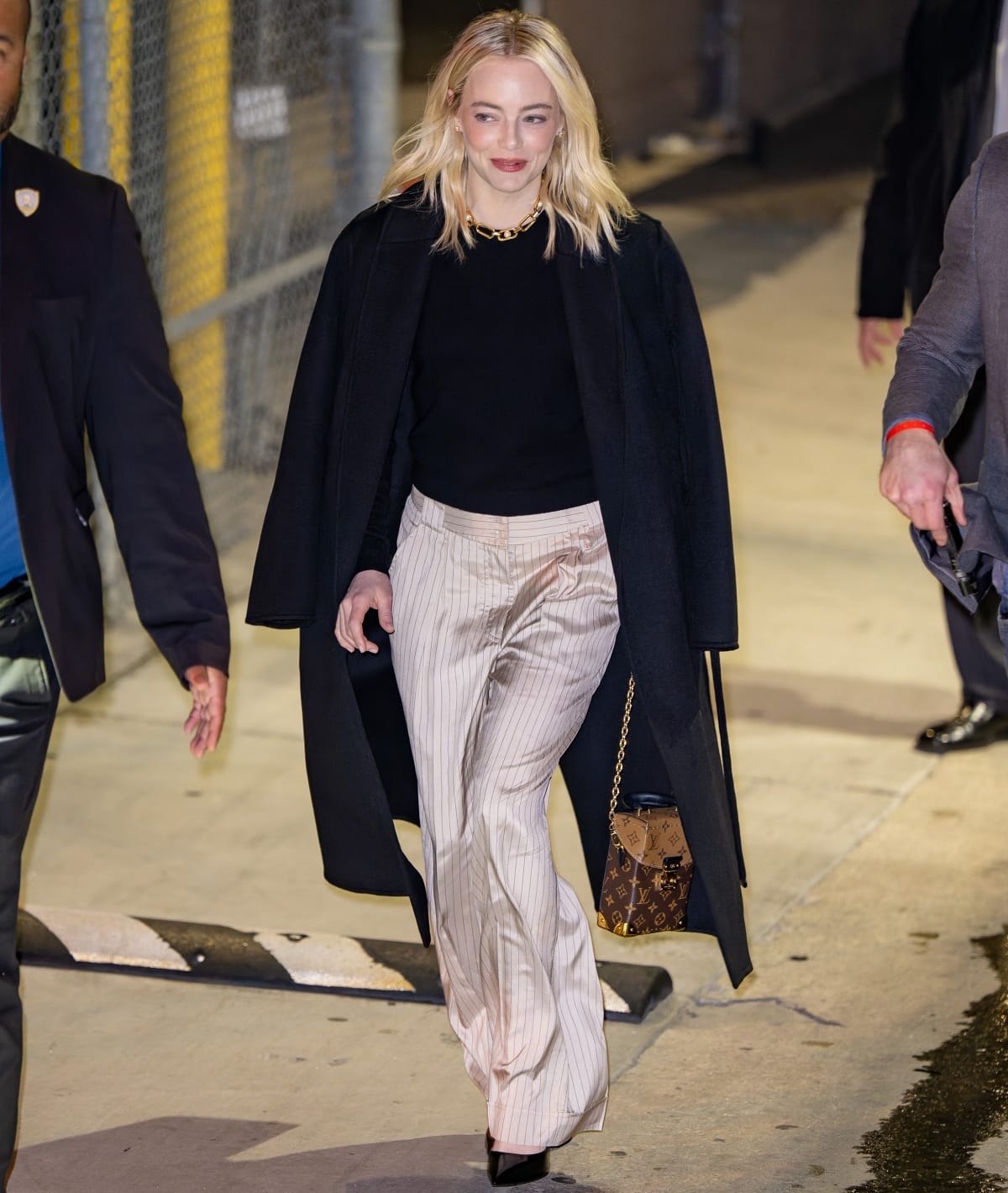 Emma Stone donning a navy overcoat with a black undershirt and champagne satin pinstripe trousers outside the Jimmy Kimmel Live! studios after her guest stint