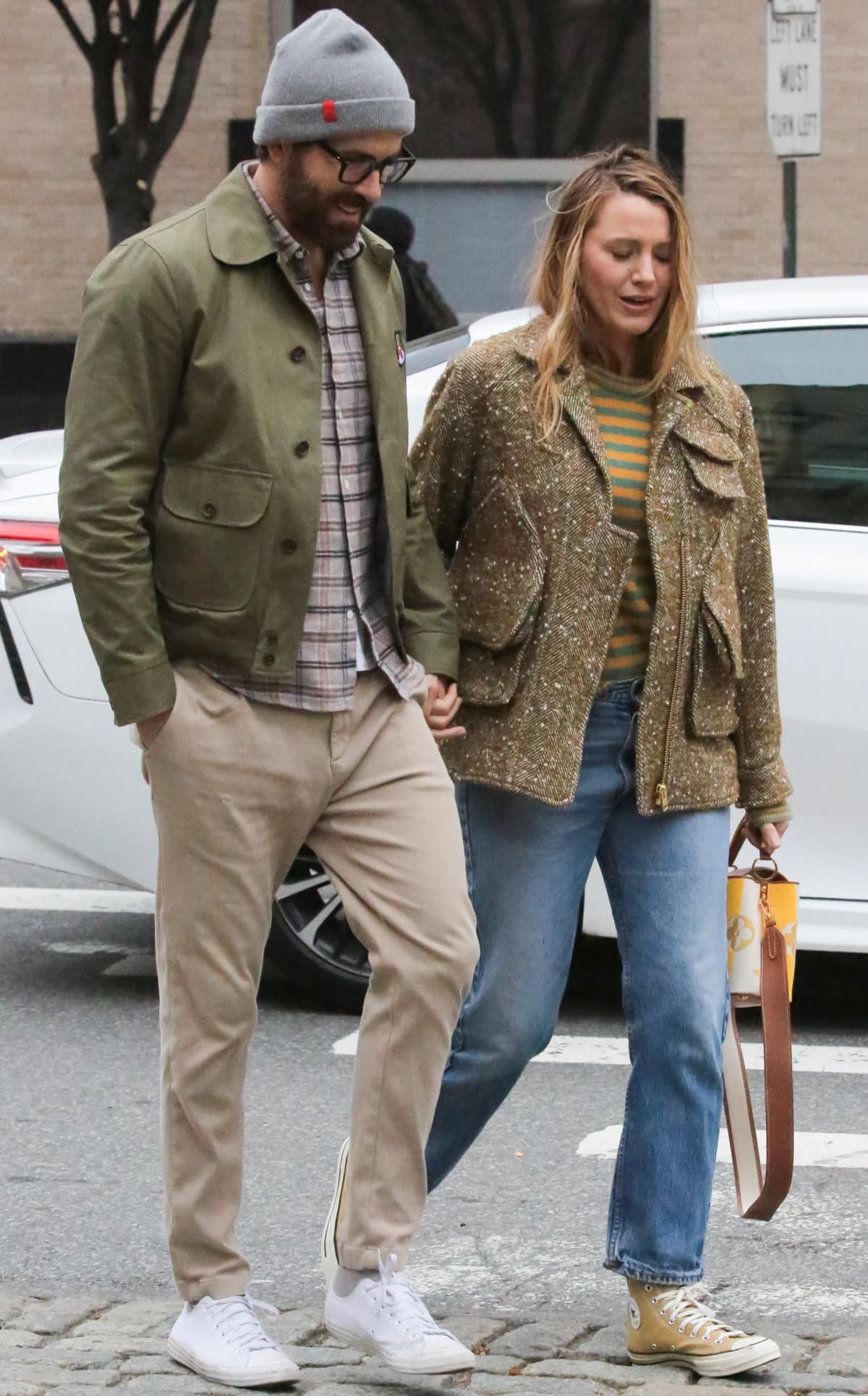 Ryan Reynolds wearing a brown plaid button-down top, beige khakis, an olive jacket, and white sneakers with a gray beanie and black-framed glasses