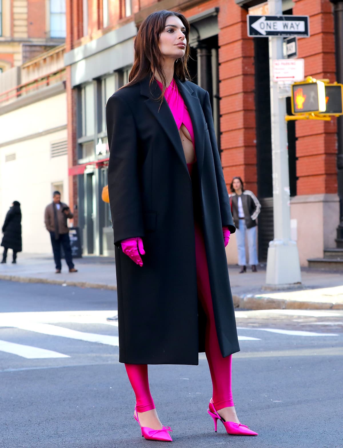 Emily Ratajkowski filming a commercial for makeup brand Maybelline in a hot pink The Andamane catsuit with a large underboob-baring cutout, matching pink gloves, and an oversized black coat on December 14, 2023