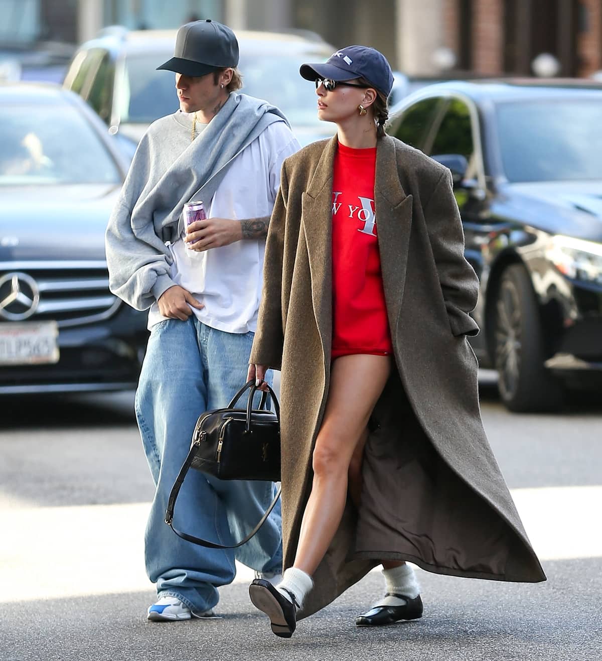 Joined by her husband, Justin Bieber, Hailey Bieber's outfit is elegantly completed by a flowing chestnut-brown coat, Sandy Liang Mary Jane Pointe shoes, and chic accessories, including gold hoop earrings and a black Nike cap