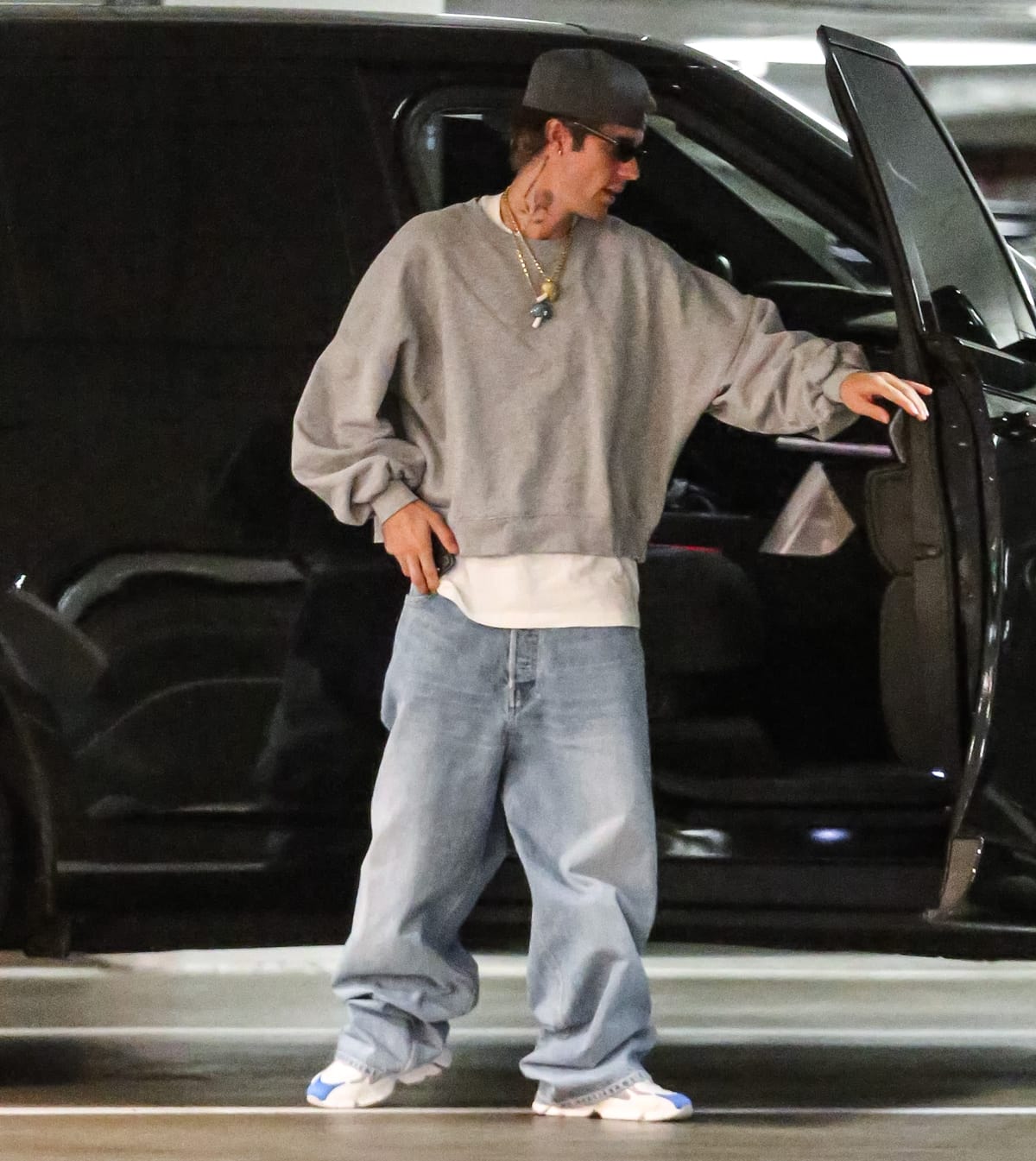 Justin Bieber pairs wide-legged jeans with a crisp white short-sleeved shirt, a casually draped grey sweatshirt, a black trucker hat, and trendy white and blue sneakers