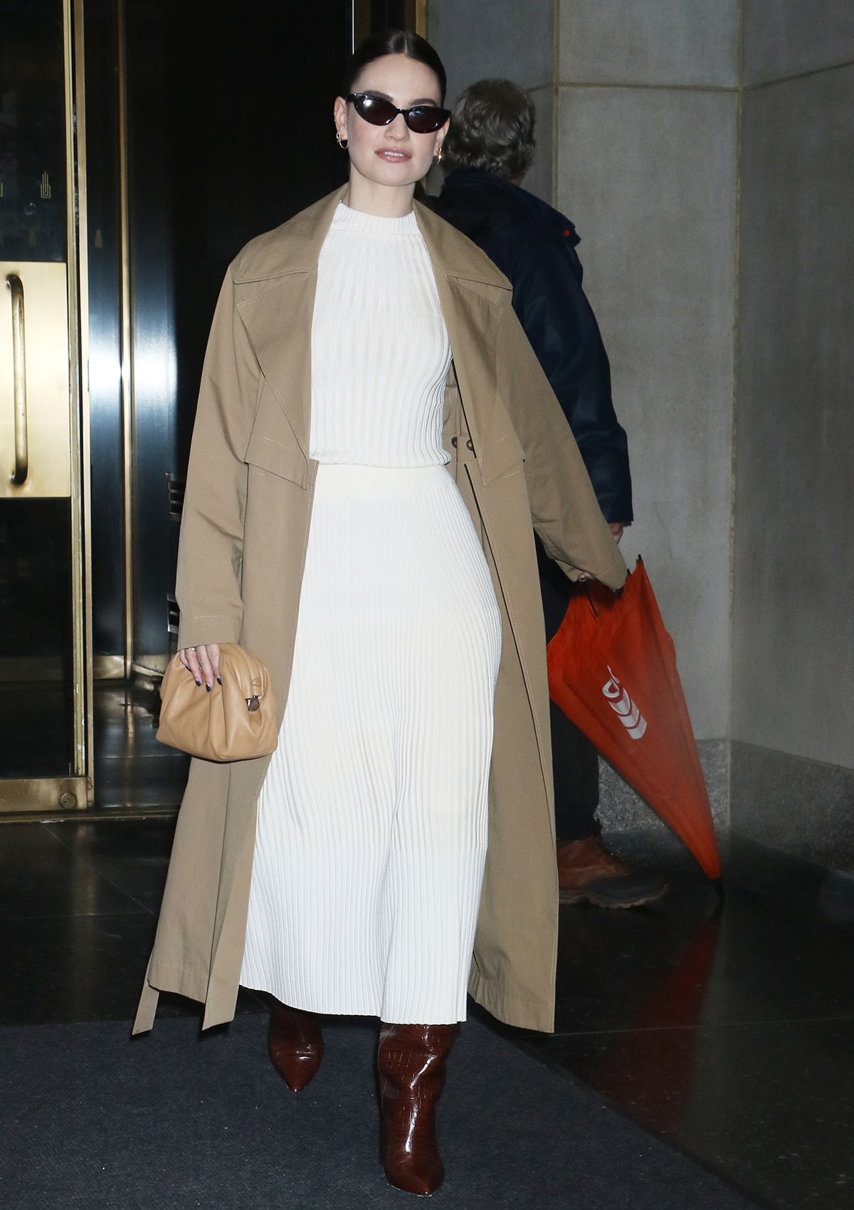 Lily James is radiant in a white Lafayette 148 ribbed sweater, a matching white sweater skirt, and a khaki convertible trench coat as she visits the Today studio to promote The Iron Claw on December 18, 2023