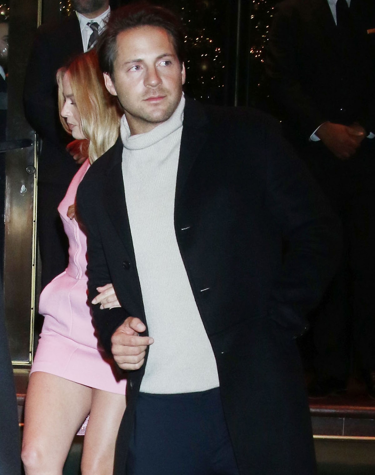 Tom Ackerley looks as dashing as Ken in a beige turtleneck, navy pants, and black peacoat