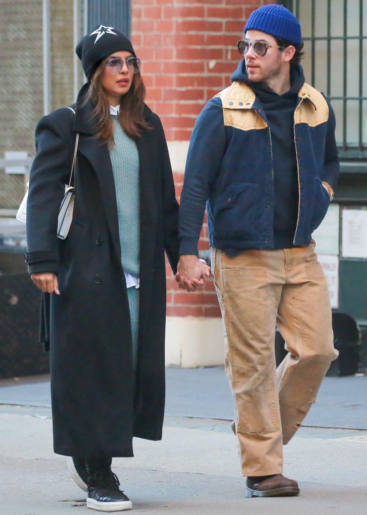 Priyanka Chopra wearing a turquoise knit dress under a long black coat with a dark blue beanie, tinted sunglasses, a white handbag, and black shoes with white soles