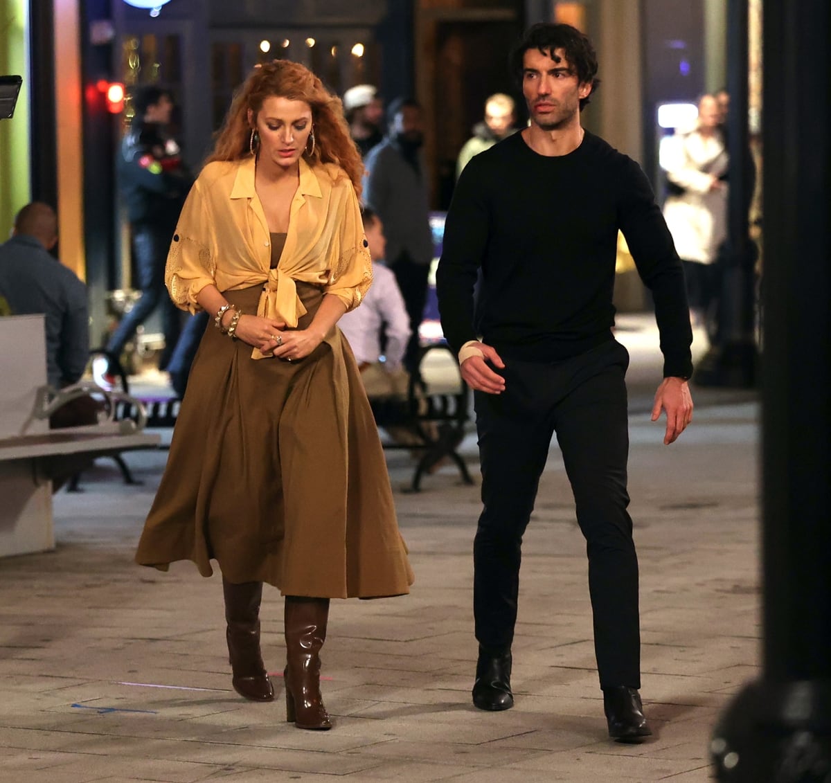 Blake Lively, in character as Lily Bloom, wears a strapless brown tea dress with a yellow shirt tied at the waist, while Justin Baldoni, as Ryle Kincaid, appears in an all-black outfit with a bandaged arm and dramatic fight scene makeup