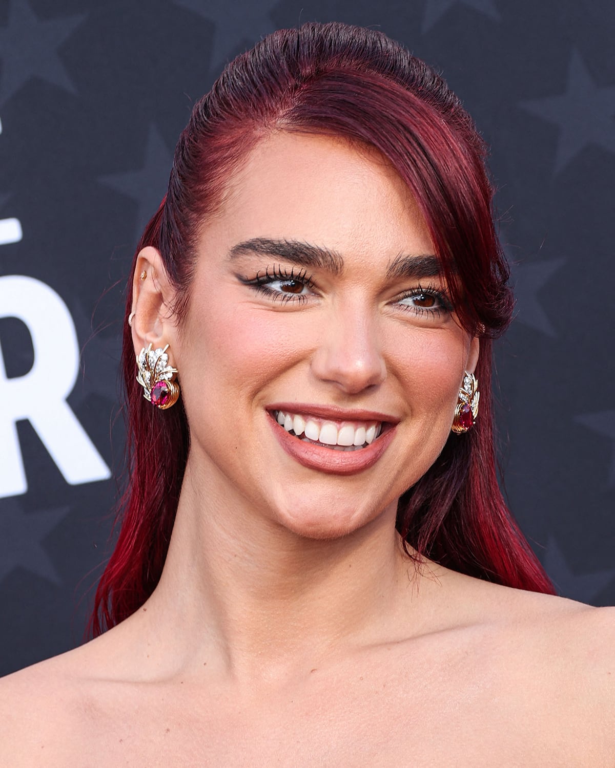 Dua Lipa styles her red hair in a half ponytail and highlights her striking features with winged eyeliner, heavy mascara, rosy blush, and lipstick