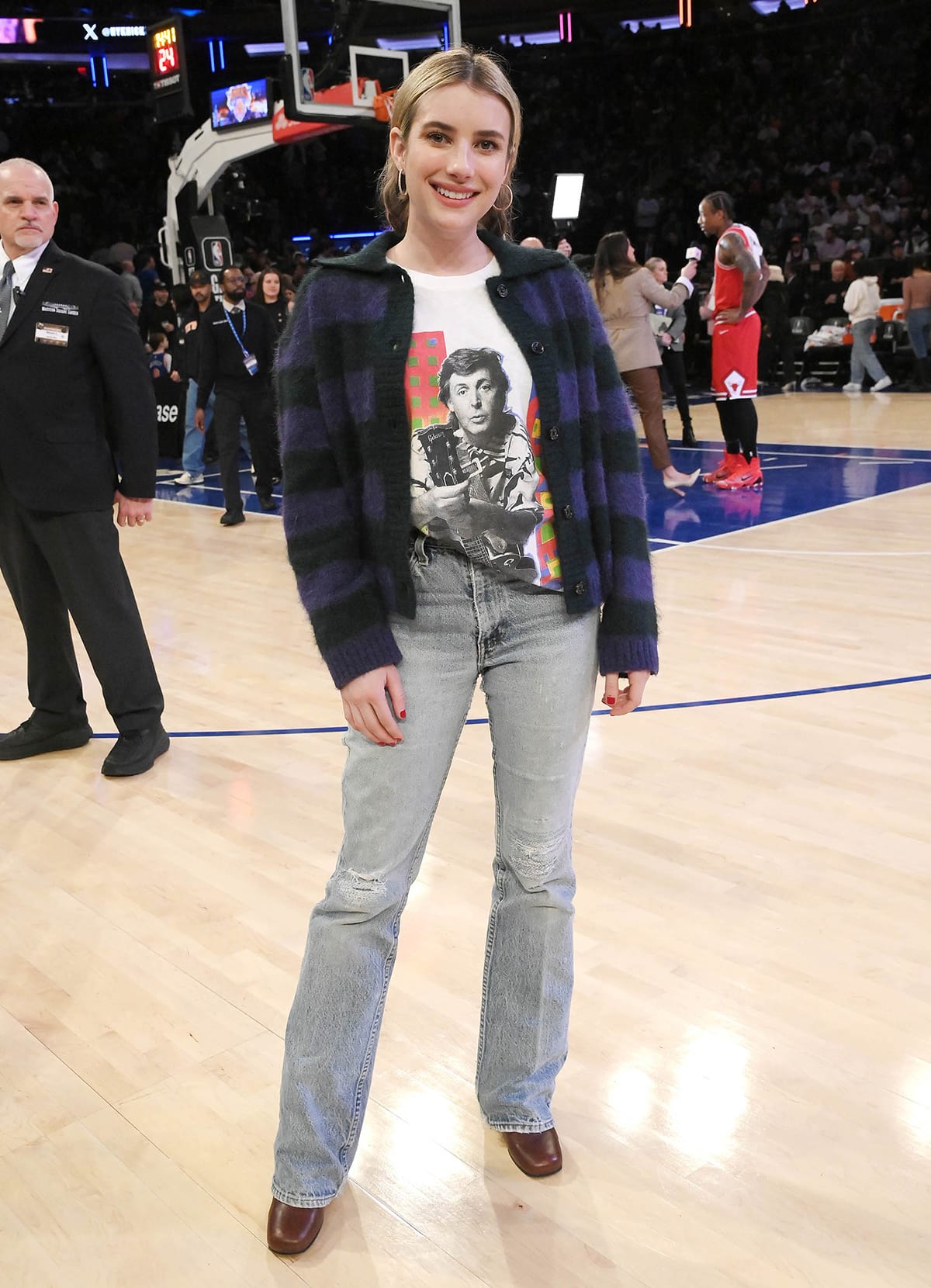 Emma Roberts looks stylish in a purple and black cardigan, vintage Paul McCartney graphic tee, and light-wash denim jeans as she sits courtside at the Madison Square Garden
