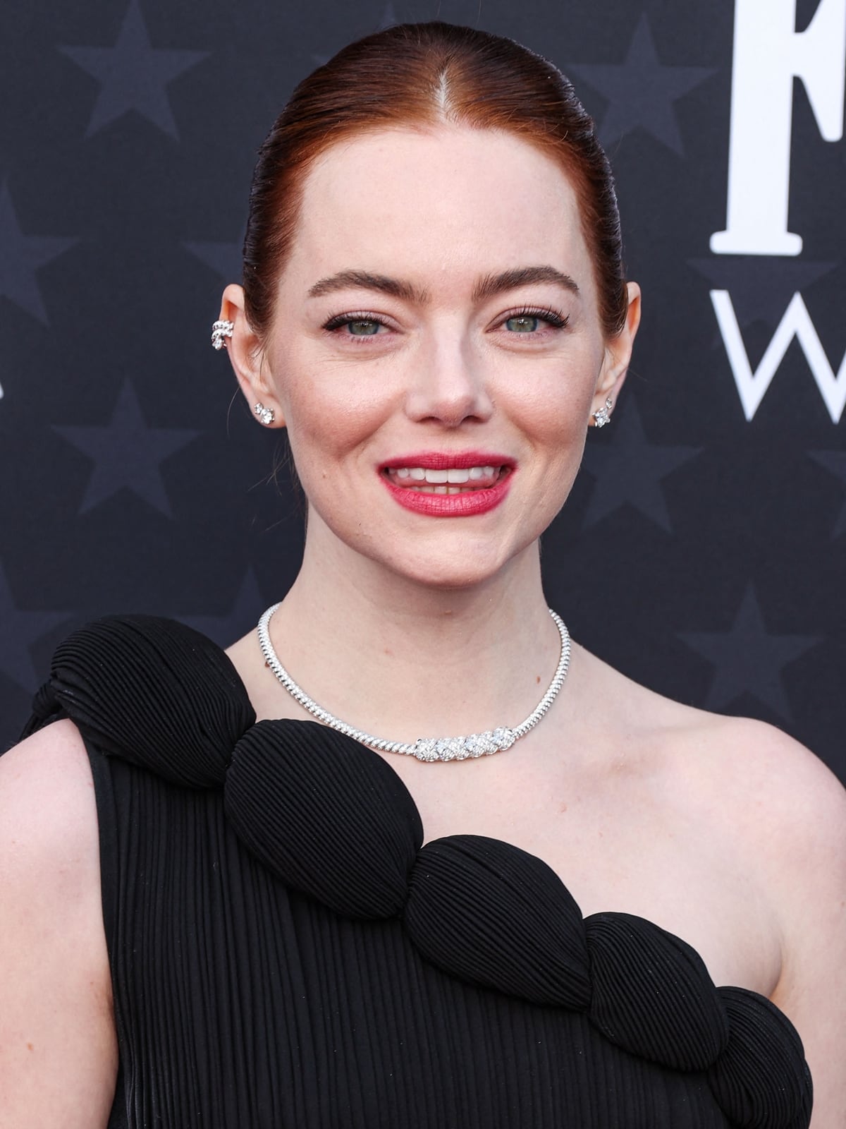 Emma Stone's vibrant red hair is styled into a sleek bun, complementing the sophisticated makeup by Rachel Goodwin, featuring warm brown eyeshadow and red sheer matte lipstick, accentuating Stone's natural beauty