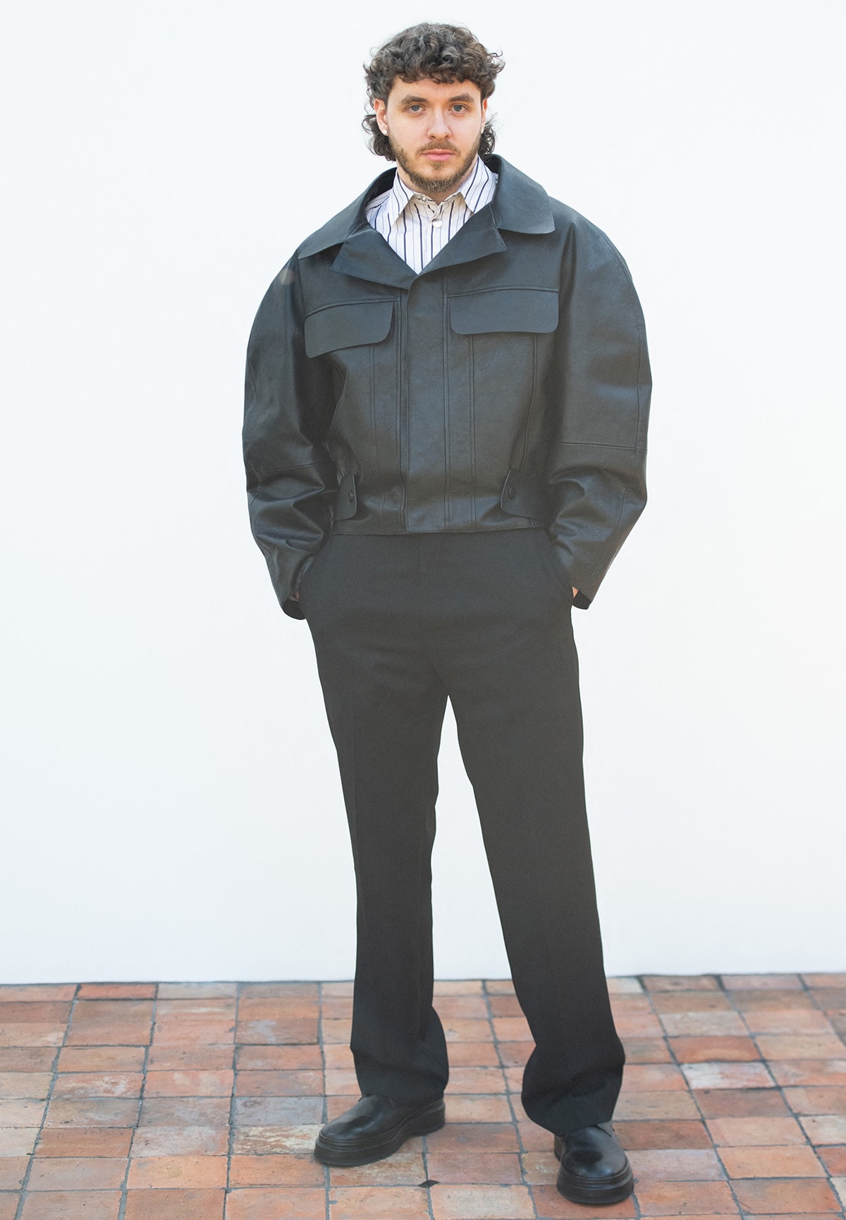 Jack Harlow looks stylish in an oversized black leather jacket, a pinstripe shirt, black trousers, and black loafers