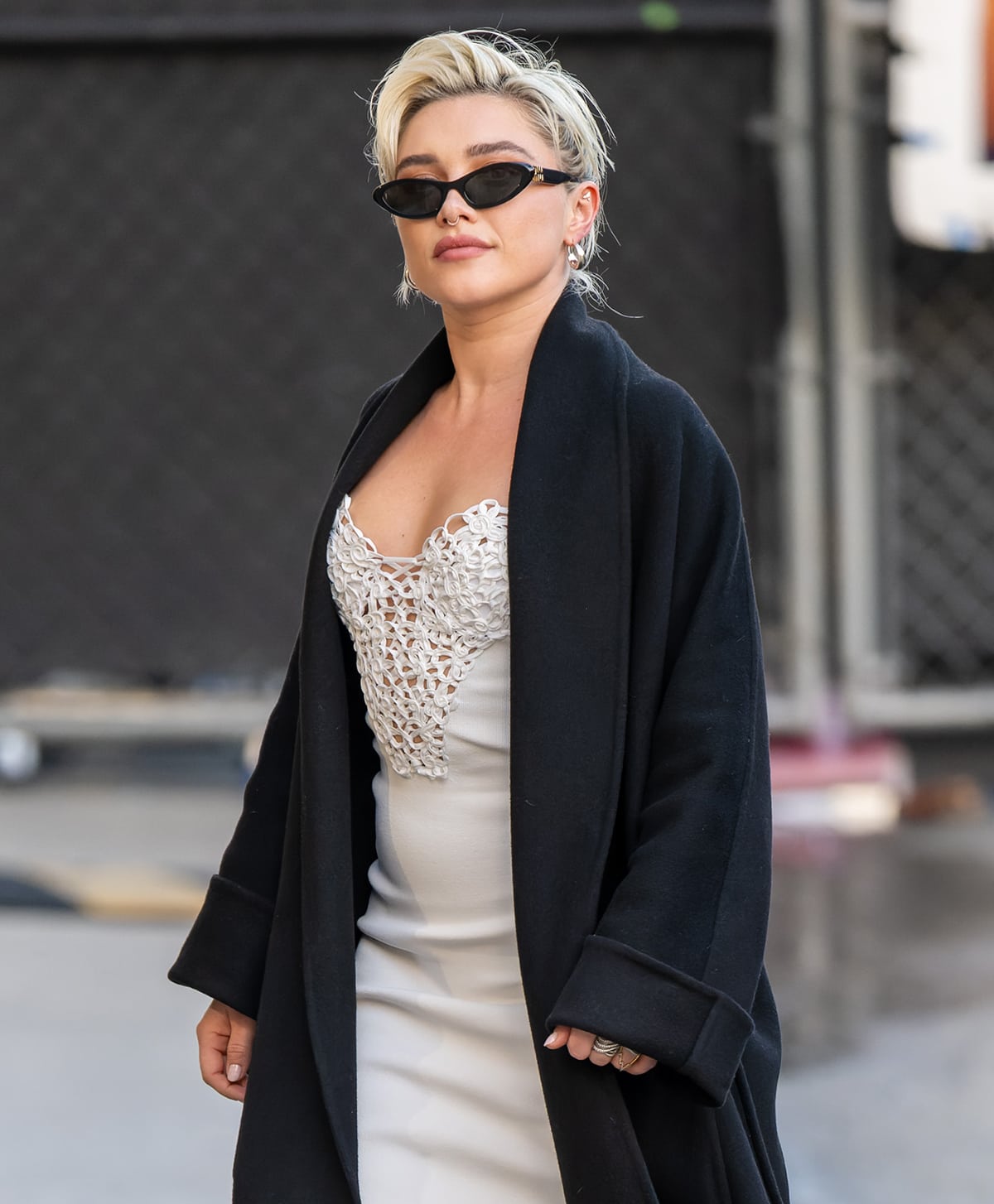 Florence Pugh adds her signature edge to her ethereal white dress by accessorizing with a black wool coat and a sleek pair of Miu Miu sunglasses