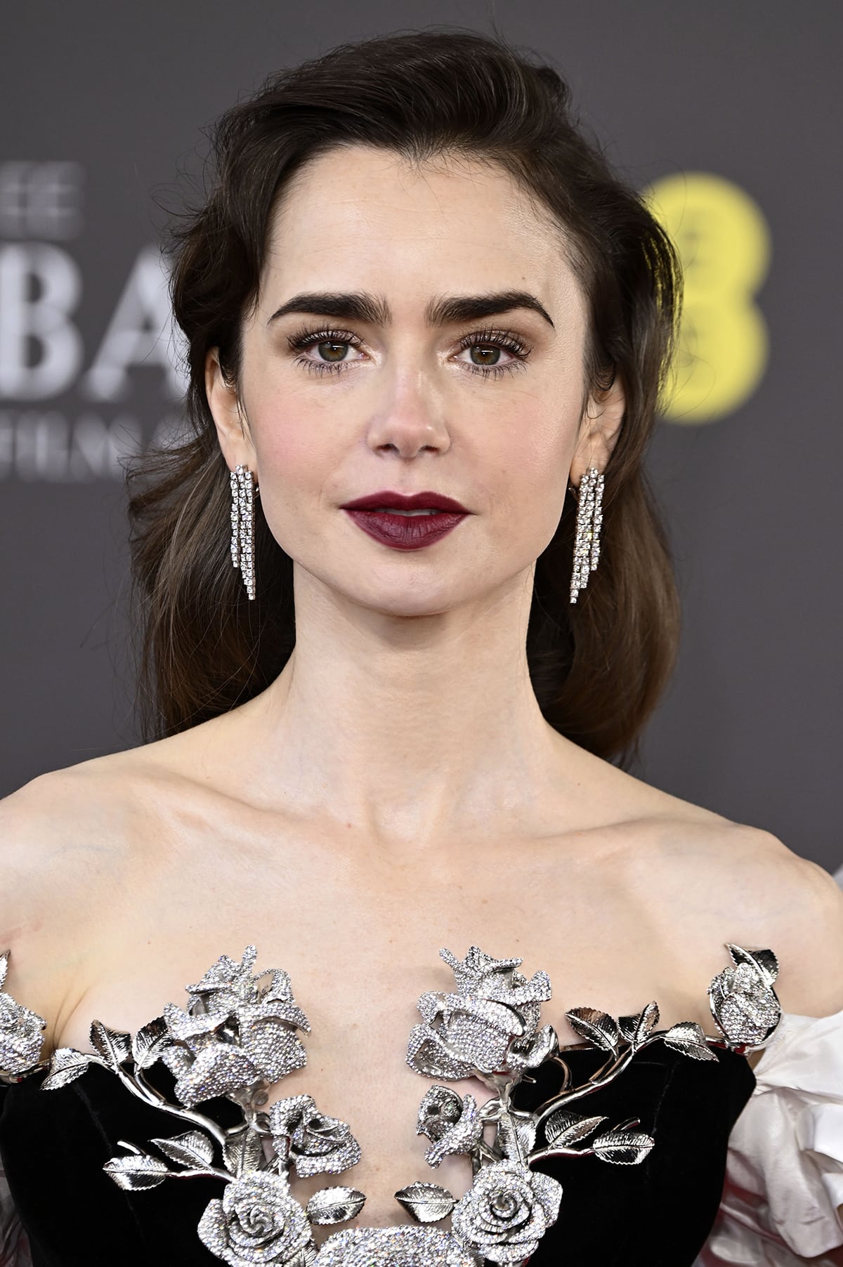 Lily Collins wears a retro side-parted hairstyle and glams up with fluttery eyelashes, shimmering smokey eyeshadow, and burgundy lipstick