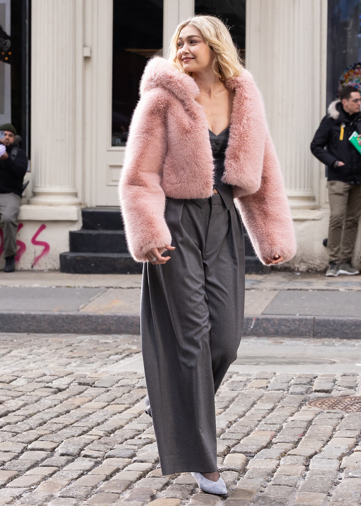 Gigi Hadid swaps her edgy business outfit for a feminine gray-and-pink ensemble with a fuzzy pink hooded jacket and a gray vest suit for her Maybelline commercial