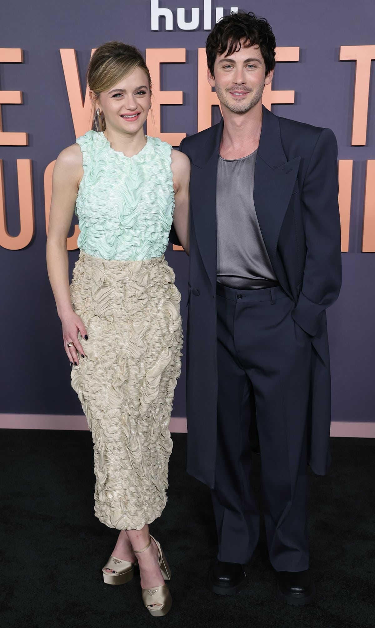Joey King and Logan Lerman shine together at the Los Angeles premiere of "We Were The Lucky Ones," with Joey in an avant-garde Prada ensemble and Logan showcasing a distinctive style in a satin tank and power shoulder overcoat