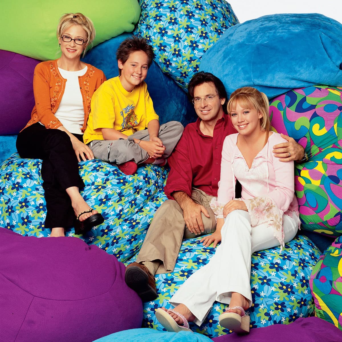 Hilary Duff, as Lizzie McGuire, rocks early 2000s teen style in a pink lace-trimmed blouse, white flared pants, and sparkly platform sandals while posing with her on-screen family