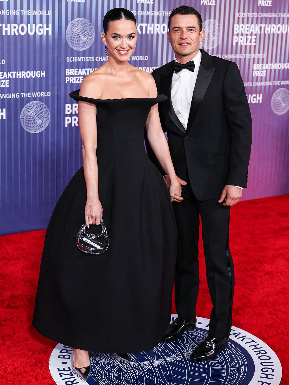 Orlando Bloom holds Katy Perry's hands while clad in a black tuxedo suit, satin-striped trousers, and black loafers