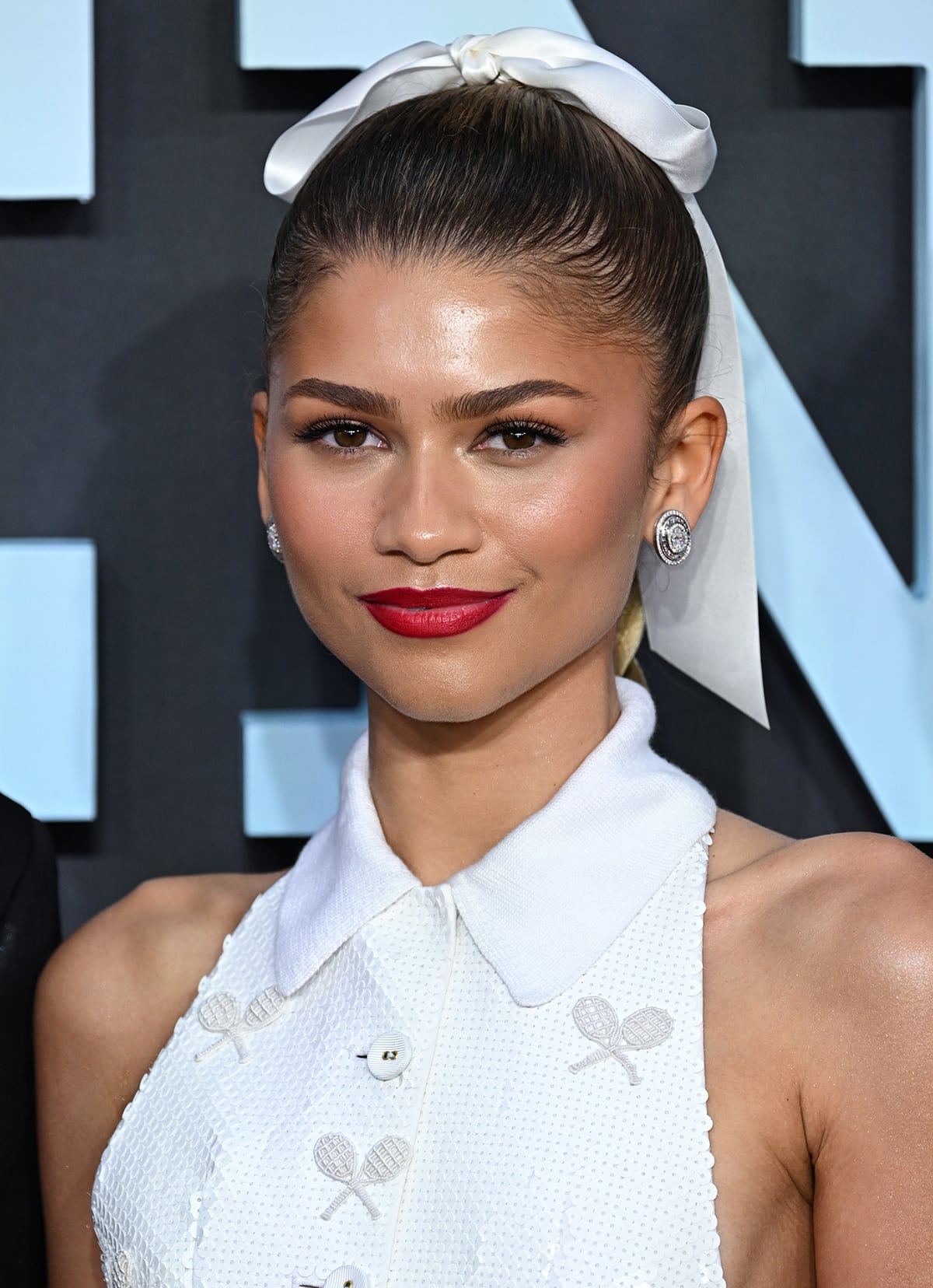 Zendaya pulls her blonde hair into a neat braided ponytail with a large white bow for a coquette touch and adds bright red lipstick to the all-white look