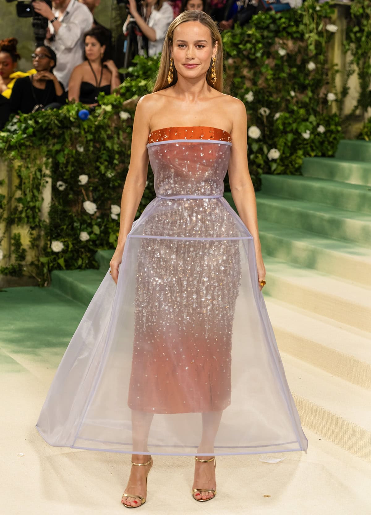 Brie Larson dazzles at the 2024 Met Gala in a Prada gown, showcasing a burnt orange satin sheath dress with a crystal-adorned bodice and a lavender hourglass overlay