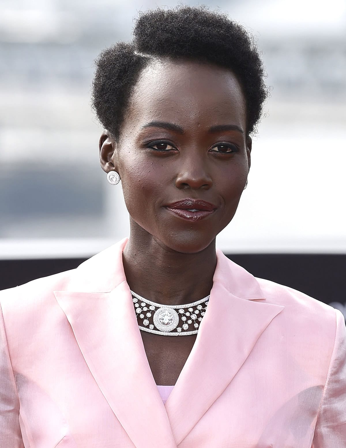 Lupita Nyong'o dazzles as she accessorizes her pink suit with De Beers Atomique Clip Earrings and Collar Necklace, inspired by the flawless symmetry of a diamond's internal molecular structure