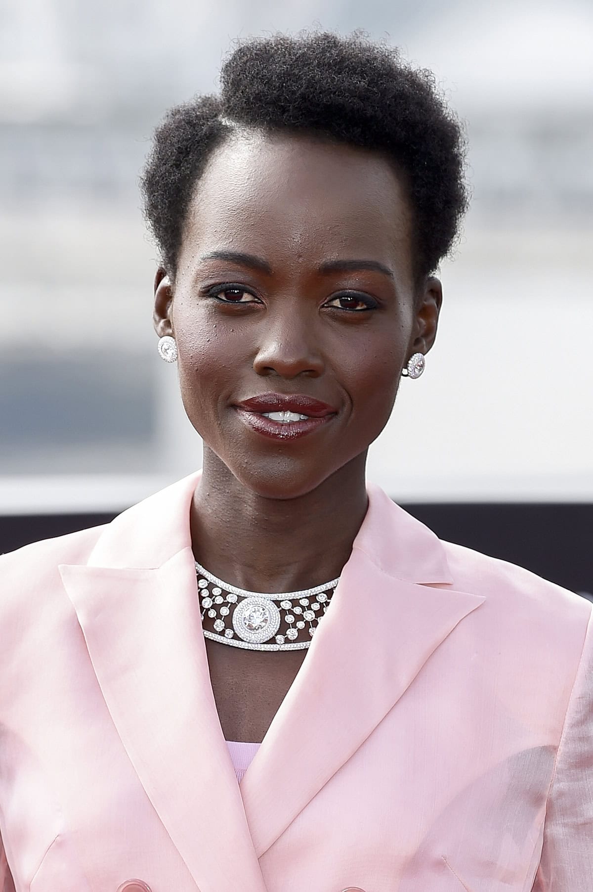 Lupita Nyong'o parts her short curly hair to the side and wears pink sorbet makeup to match her outfit