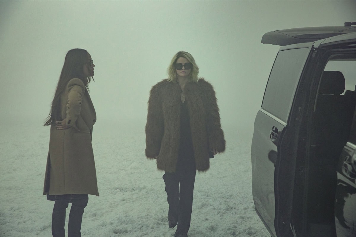 Selena Gomez, as Jessi, exudes quiet power in a Saint Laurent fur coat and heeled boots while walking through the snowy landscape beside Zoe Saldaña’s Rita Moro Castro in Emilia Pérez