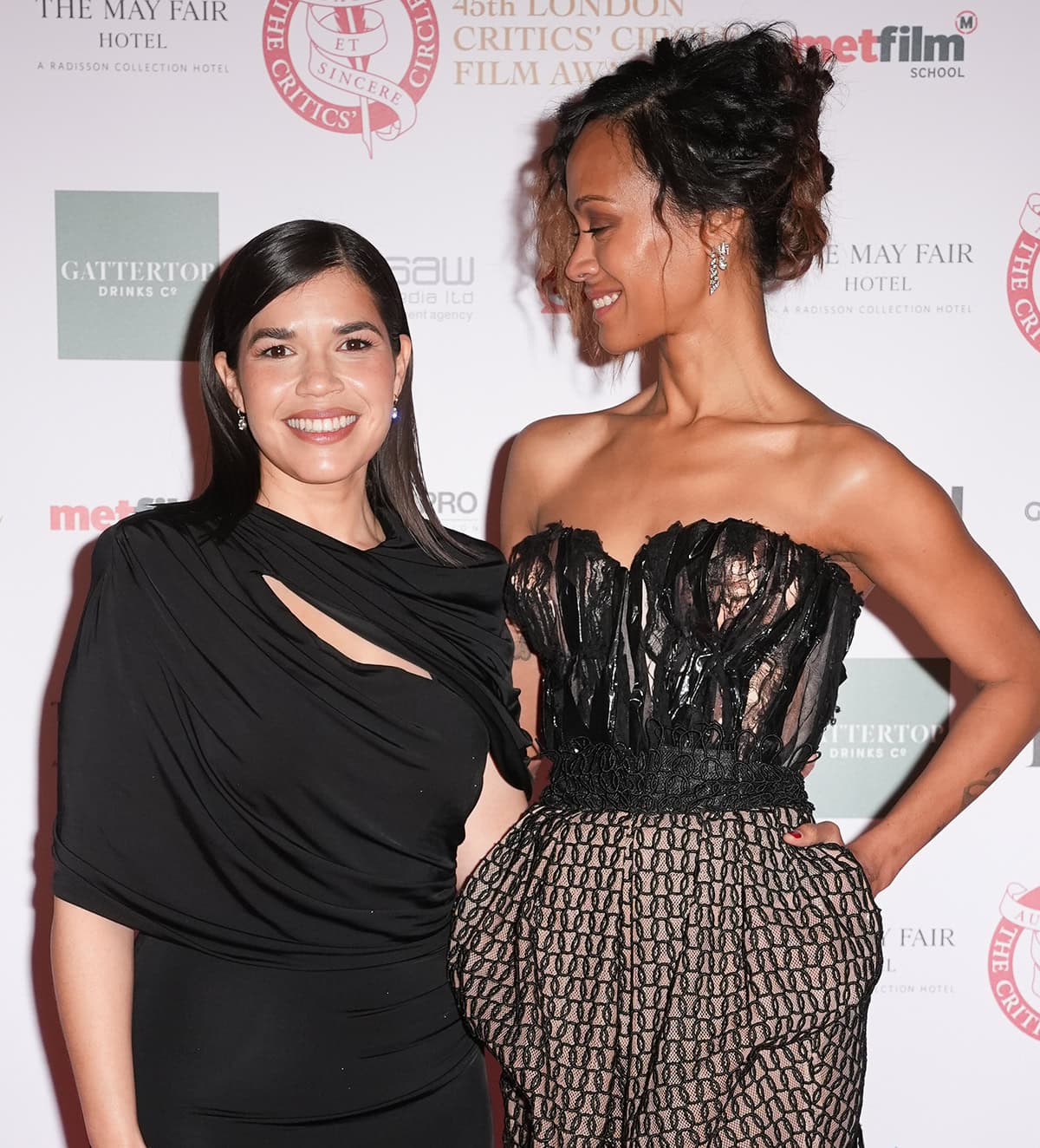 Zoe Saldaña, towering over America Ferrera, exudes elegance in her avant-garde Maison Margiela dress, while America stuns in a sleek black gown; Zoe’s two-toned curly tresses styled in a messy updo complete her edgy look