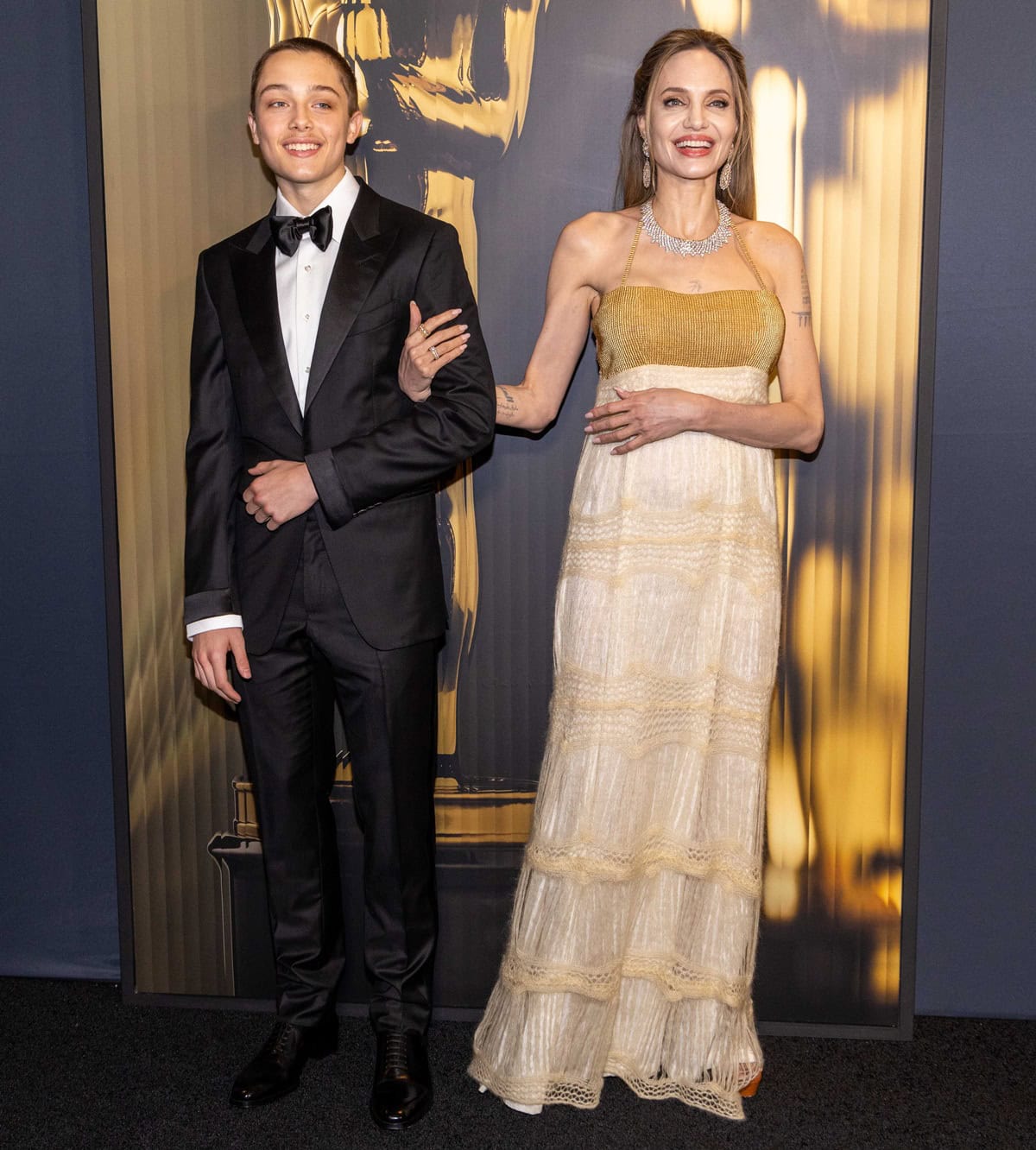 Angelina Jolie embraced her signature quiet-luxury aesthetic at the 15th Governors Awards in a shimmering archival gown with a gold-beaded bodice, finishing the look with discreet neutral open-toe sandals that subtly added height beneath the flowing hem, while Knox Jolie-Pitt complemented the formal mood in a classic black tuxedo and polished patent leather dress shoes