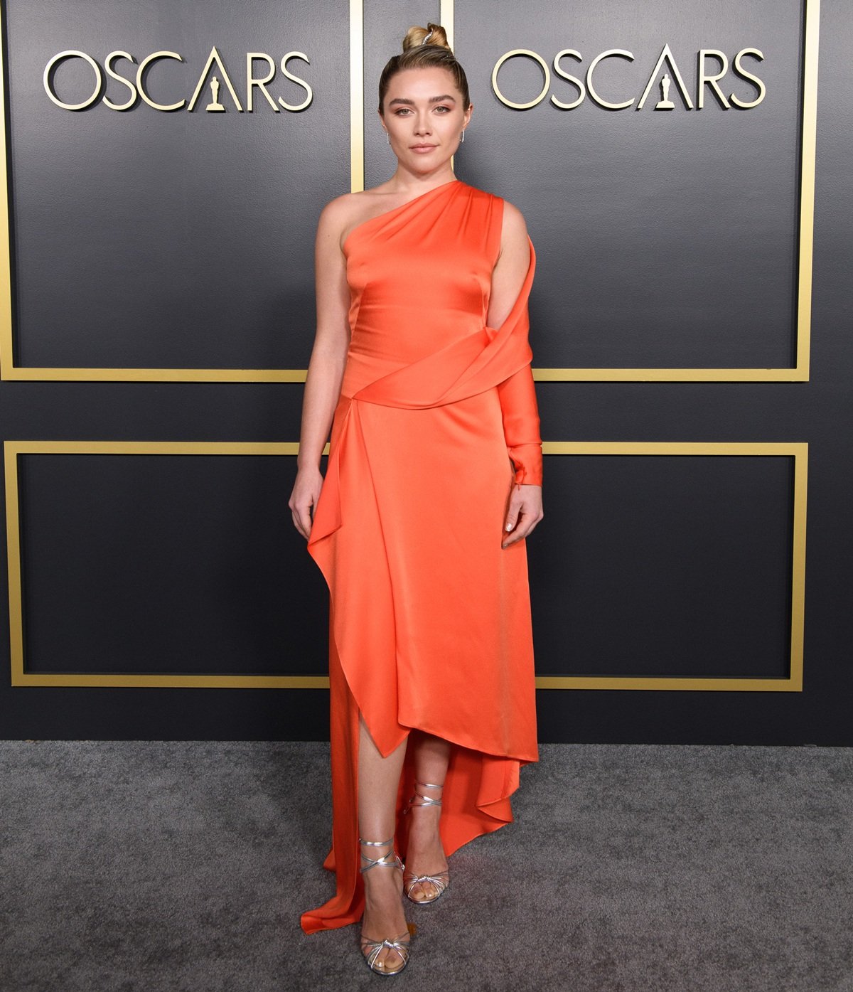 Florence Pugh stuns in a bold tangerine one-shoulder silk dress by Monse, paired with strappy metallic Jimmy Choo sandals and diamond safety pin earrings by Anita Ko at the 2020 Oscar Nominee Luncheon