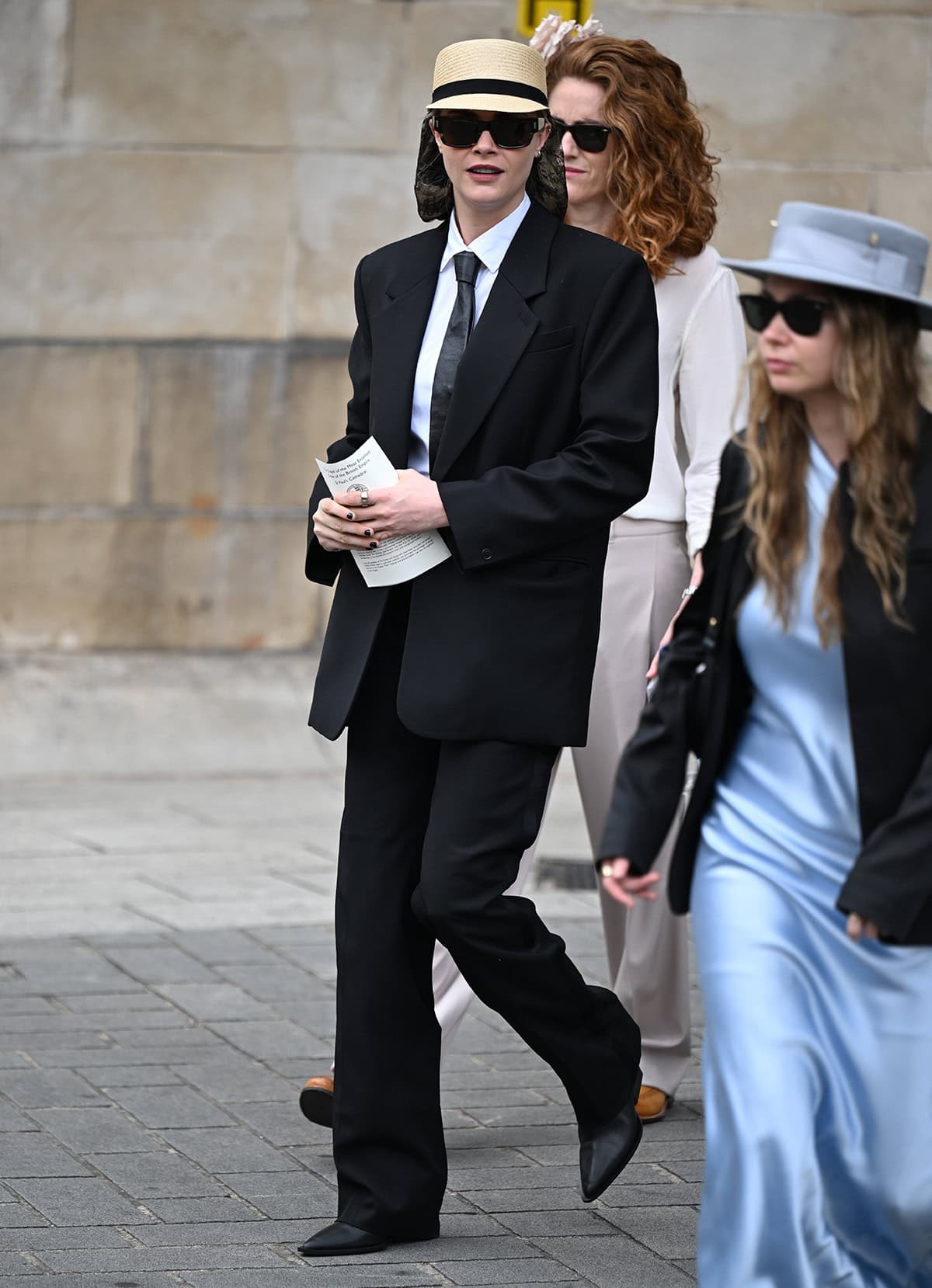 Cara Delevingne brings her signature androgynous style to Mel B's wedding, wearing a black Camilla and Marc suit, a white shirt, a necktie, and pointed black shoes