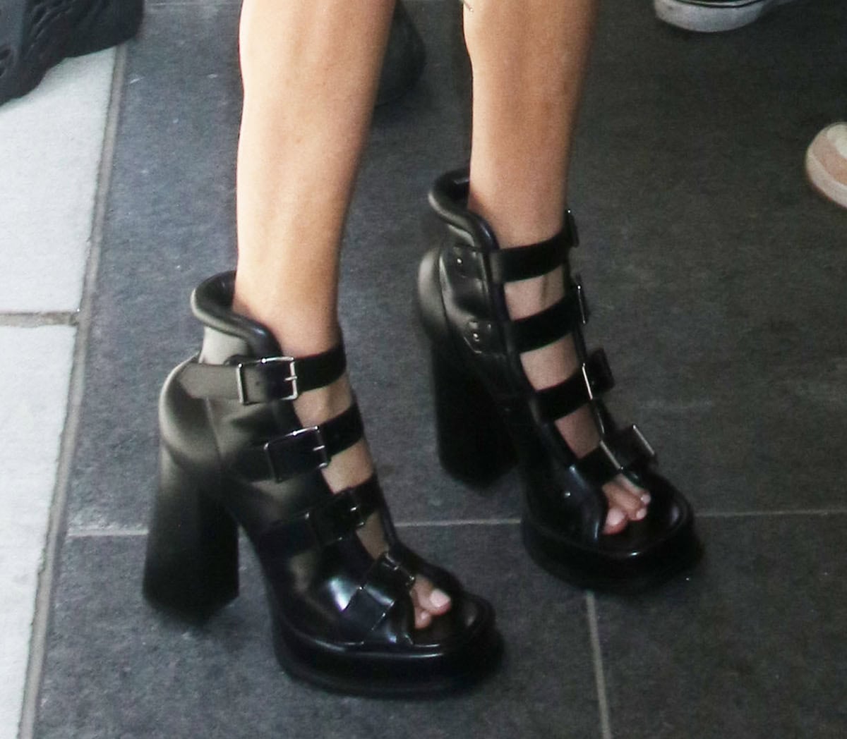 Charlize Theron embraces a bold and edgy aesthetic with Alexander McQueen's Caged sandals featuring four thick buckled straps, chunky platforms, bombe heel counters, and slanted block heels