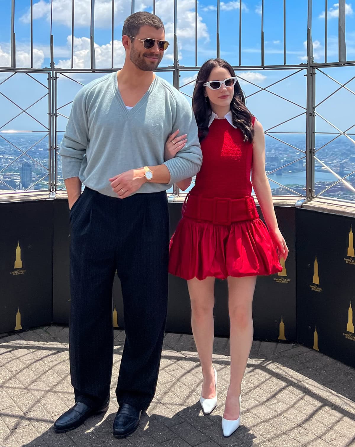 Rachel Brosnahan looks youthful in a red sleeveless Patou dress with a white collar peeking from the neckline, large buckled belt detail, and a bubble skirt