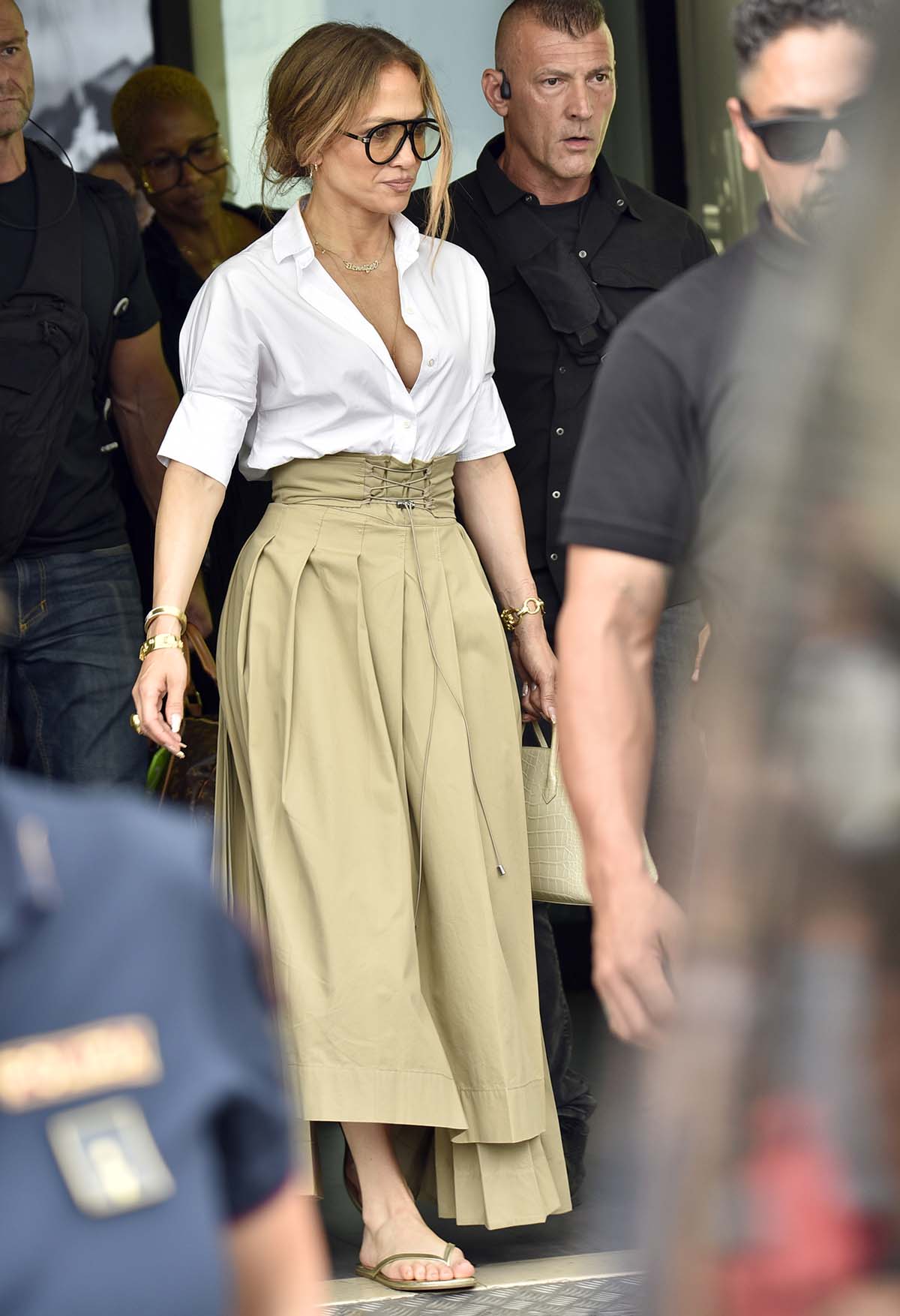Jennifer Lopez's summer-chic airport outfit includes a white Dior cotton poplin shirt and a khaki skirt featuring a lace corset waist, sharp pleats, and an asymmetrical hemline
