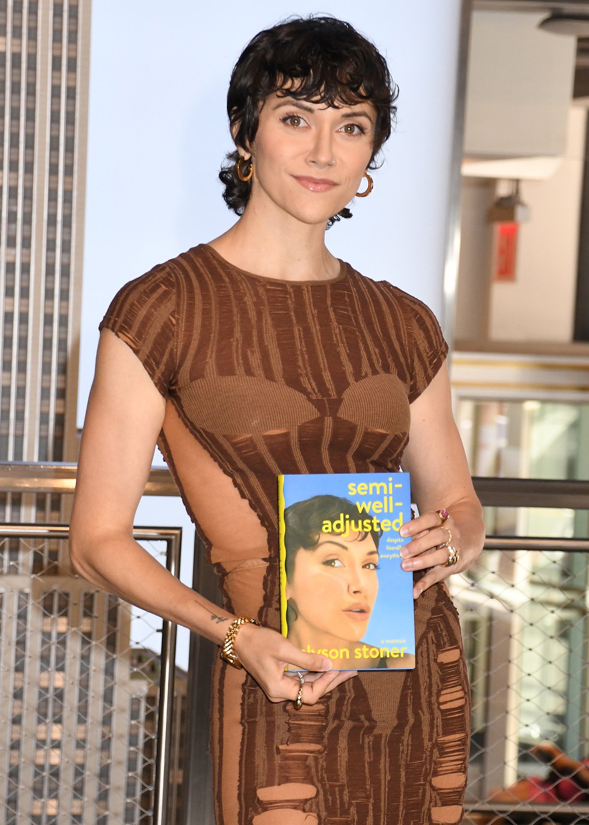 Alyson Stoner styles their brunette pixie in messy curls, wears neutral makeup, and accessorizes with gold pieces from Sif Jakobs Jewellery