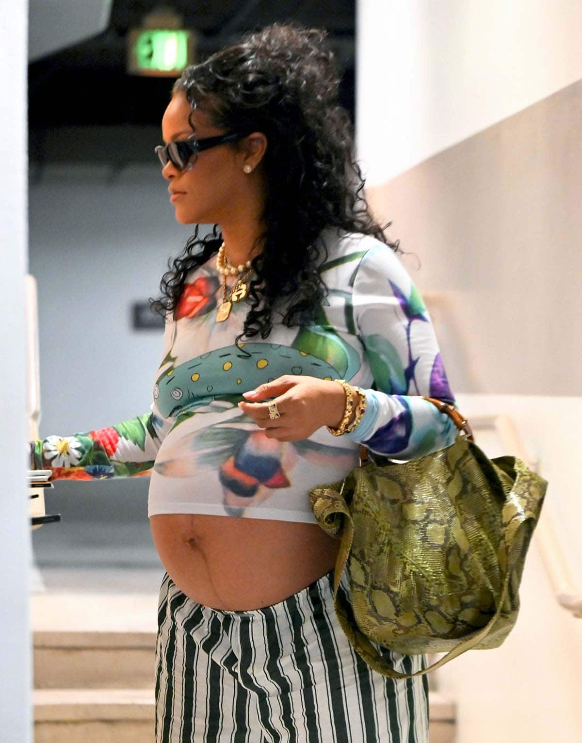 Rihanna styles her look with a mix of chunky gold bling, including a Renato Cipullo “R” pendant necklace, a Marie Lichtenberg gold scapular necklace, and rings by XIV Karats, and carries her essentials in a green python bag from Gucci by Tom Ford