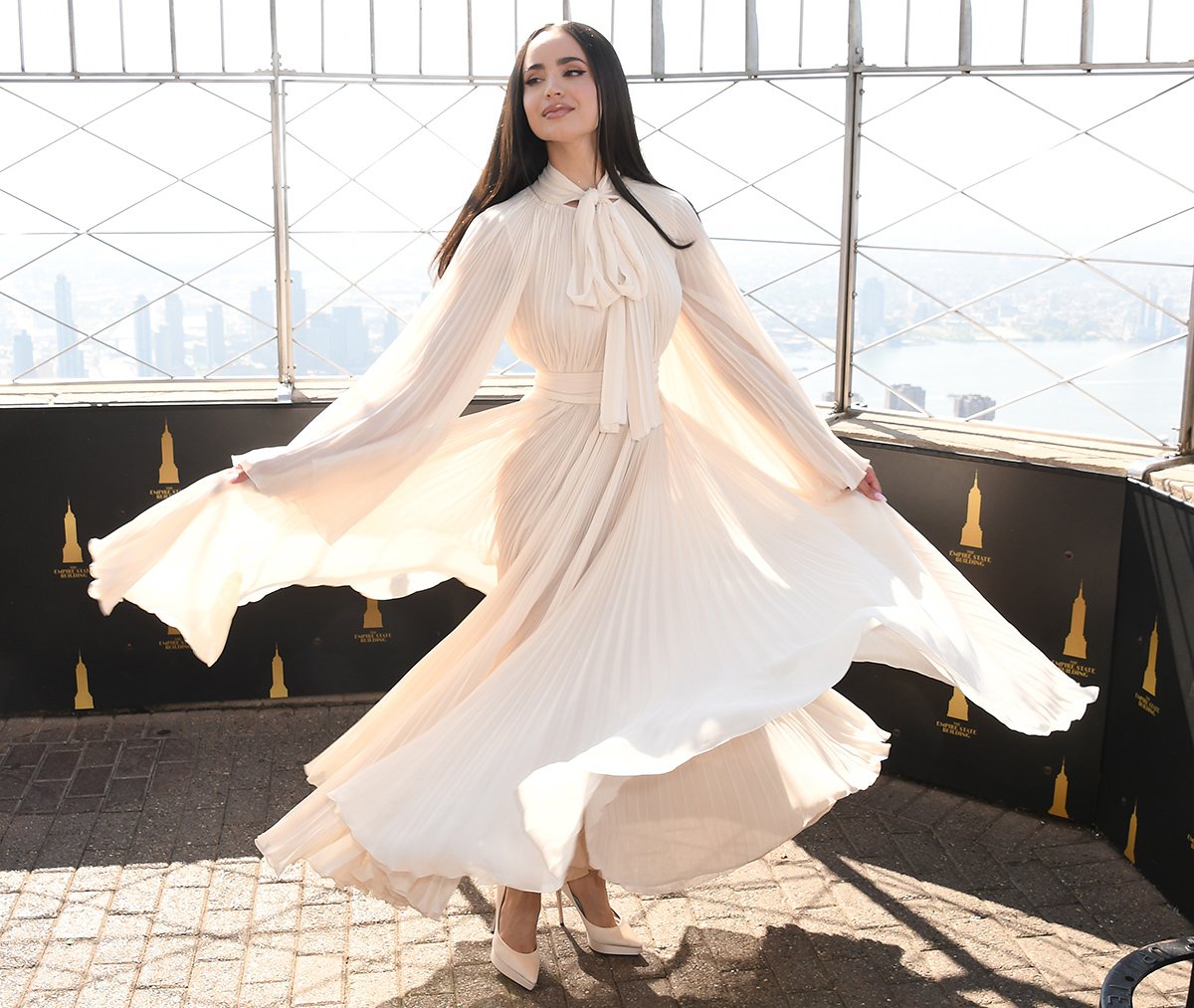 Sofia Carson twirls in an airy ivory chiffon Dolce & Gabbana gown, featuring a pussy bow, a tonal belt, and delicate pleats, at the Empire State Building