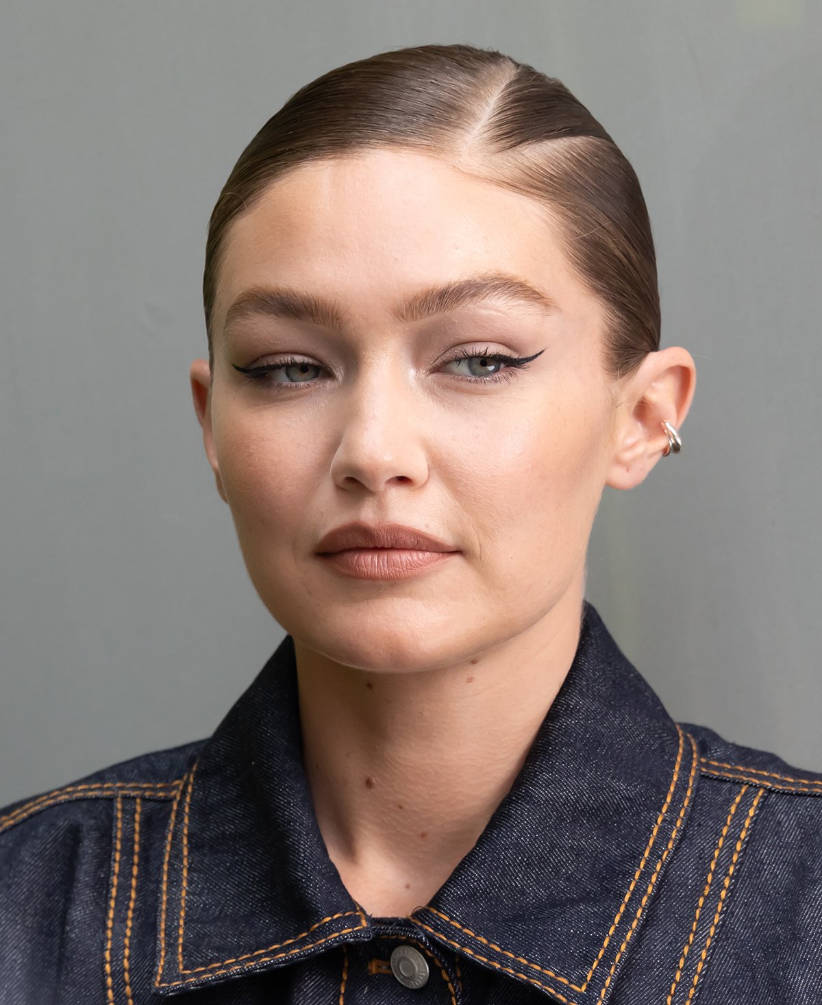 Gigi Hadid styles her hair in a slick ponytail with a V-shaped parting and highlights her features with winged eyeliner, nude blush, and lipstick