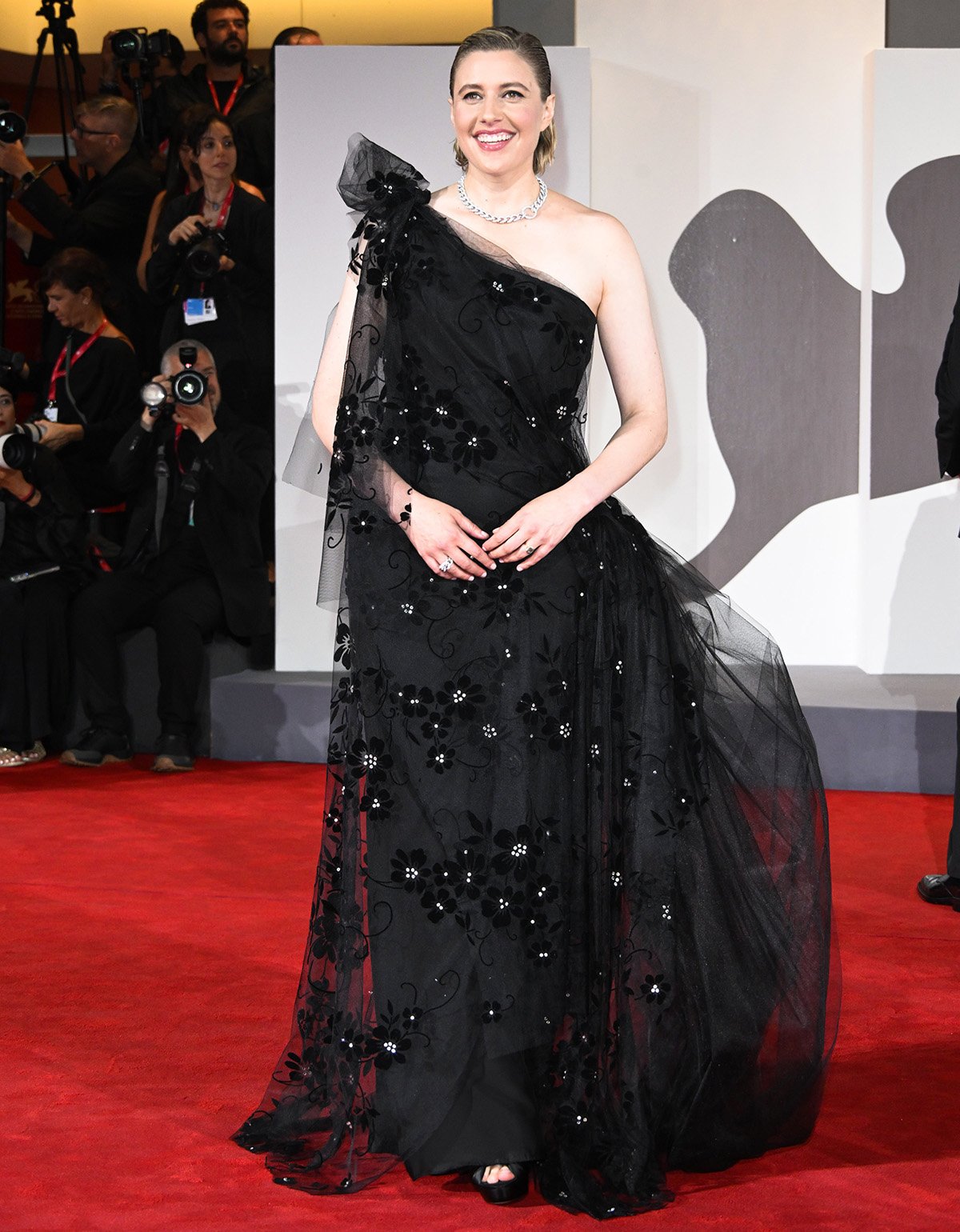 Greta Gerwig channels romantic elegance in a black tulle one-shoulder gown by Rodarte featuring crystal-adorned velvet flowers and knot accents at the shoulder and waist