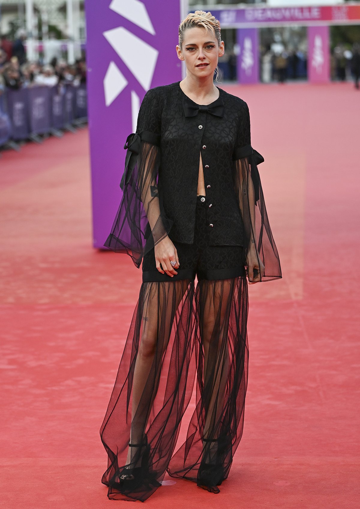 Kristen Stewart balances edge and elegance at the closing ceremony of the 51st Deauville American Film Festival, wearing a black Chanel blouse and shorts, featuring tonal monogram embroidery, black satin bows, and sheer panels cascading from the cuffs and hem