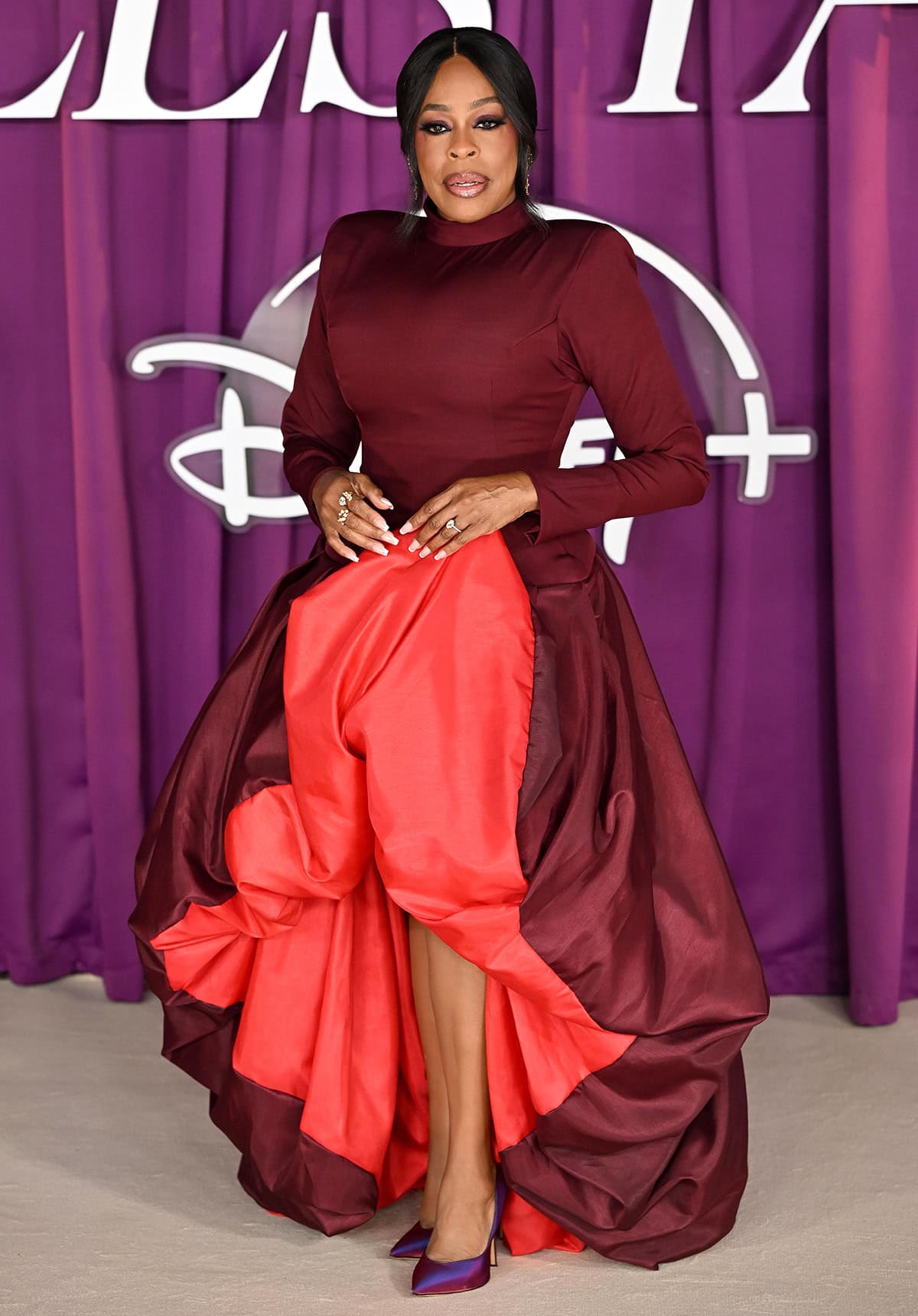 Niecy Nash-Betts brings glamour to the red carpet in a two-toned MSO Lagos gown with a deep maroon bodice and a voluminous high-low skirt with a bright red lining