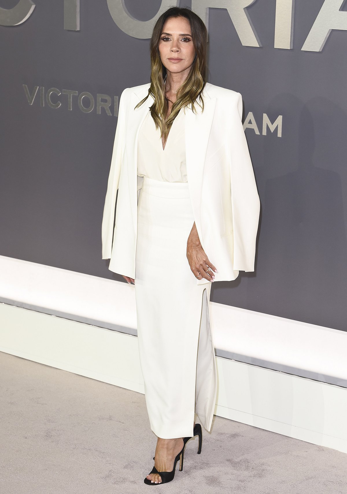 Victoria Beckham is the epitome of modern elegance in an all-white ensemble from her eponymous collection, featuring a draped ivory blouse with a plunging neckline, a white midi skirt with a side slit, and a white blazer draped over her shoulders