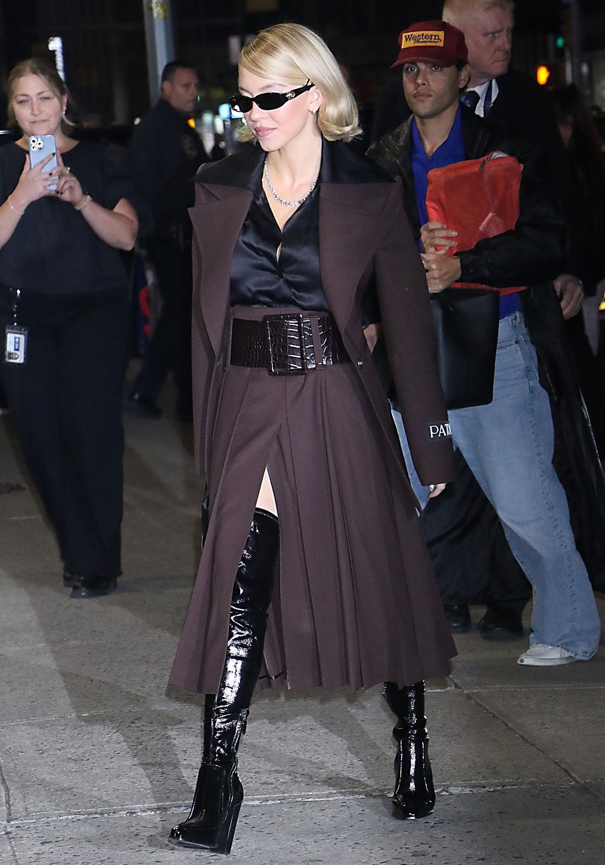 Sydney Sweeney showcases a modern, power look by pairing her black thigh-high boots with a plum Patou blazer and a matching plum pleated midi skirt with a croc-embossed leather belt