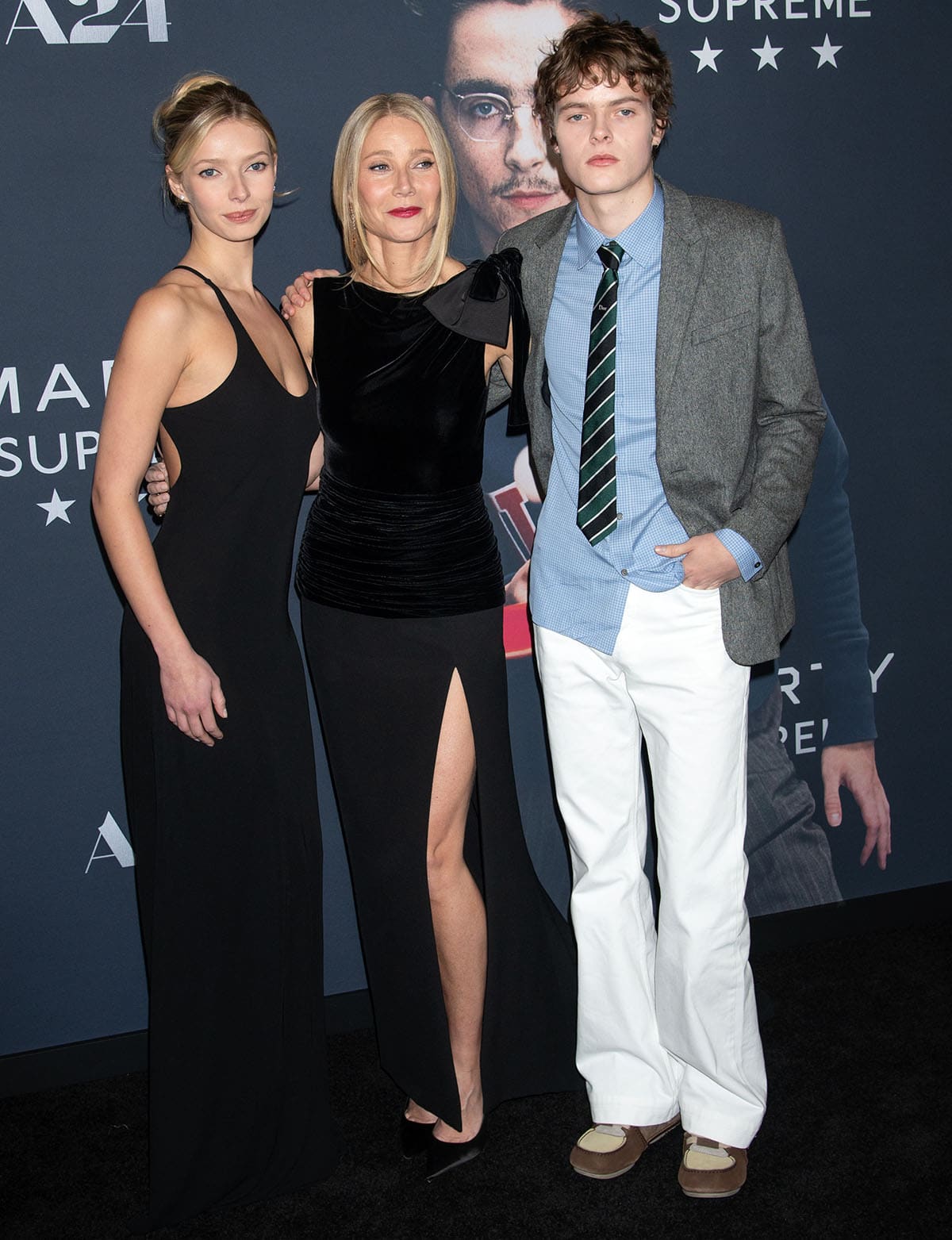 Gwyneth Paltrow, with her kids Apple and Moses Martin, embraces understated luxury in a black Valentino dress featuring a sleeveless velvet bodice with a large bow on one shoulder, a drop waist, and a thigh-high slit