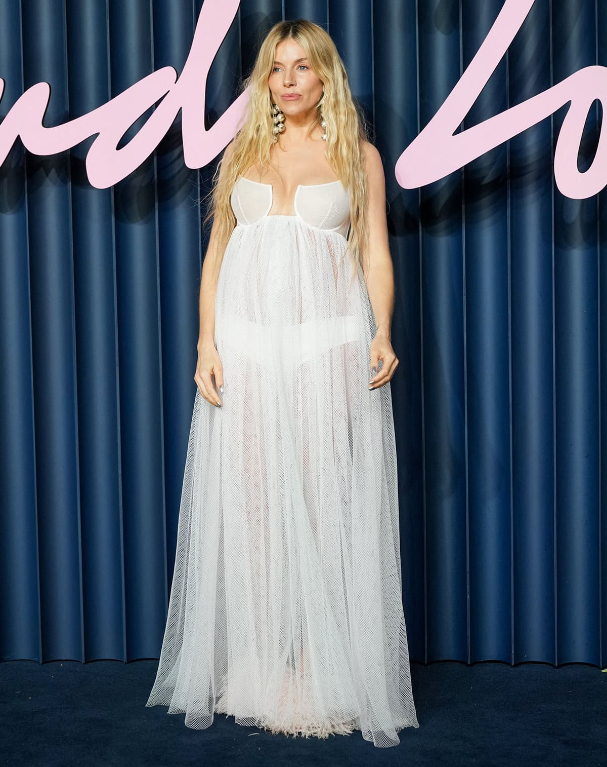 Sienna Miller reveals her baby bump in a sheer white gown made from a loose diamond-net fabric, with bra cups and a cutaway at the plunging neckline