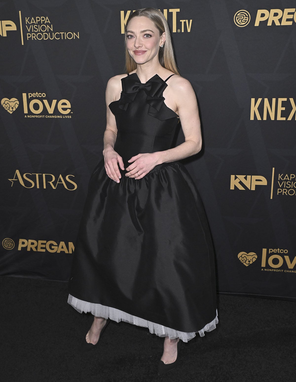 Amanda Seyfried embraces her feminine aesthetic in a black Shushu/Tong dress featuring a halter neckline with a sculptural bow accent, a voluminous skirt, and a white tiered tulle underlayer