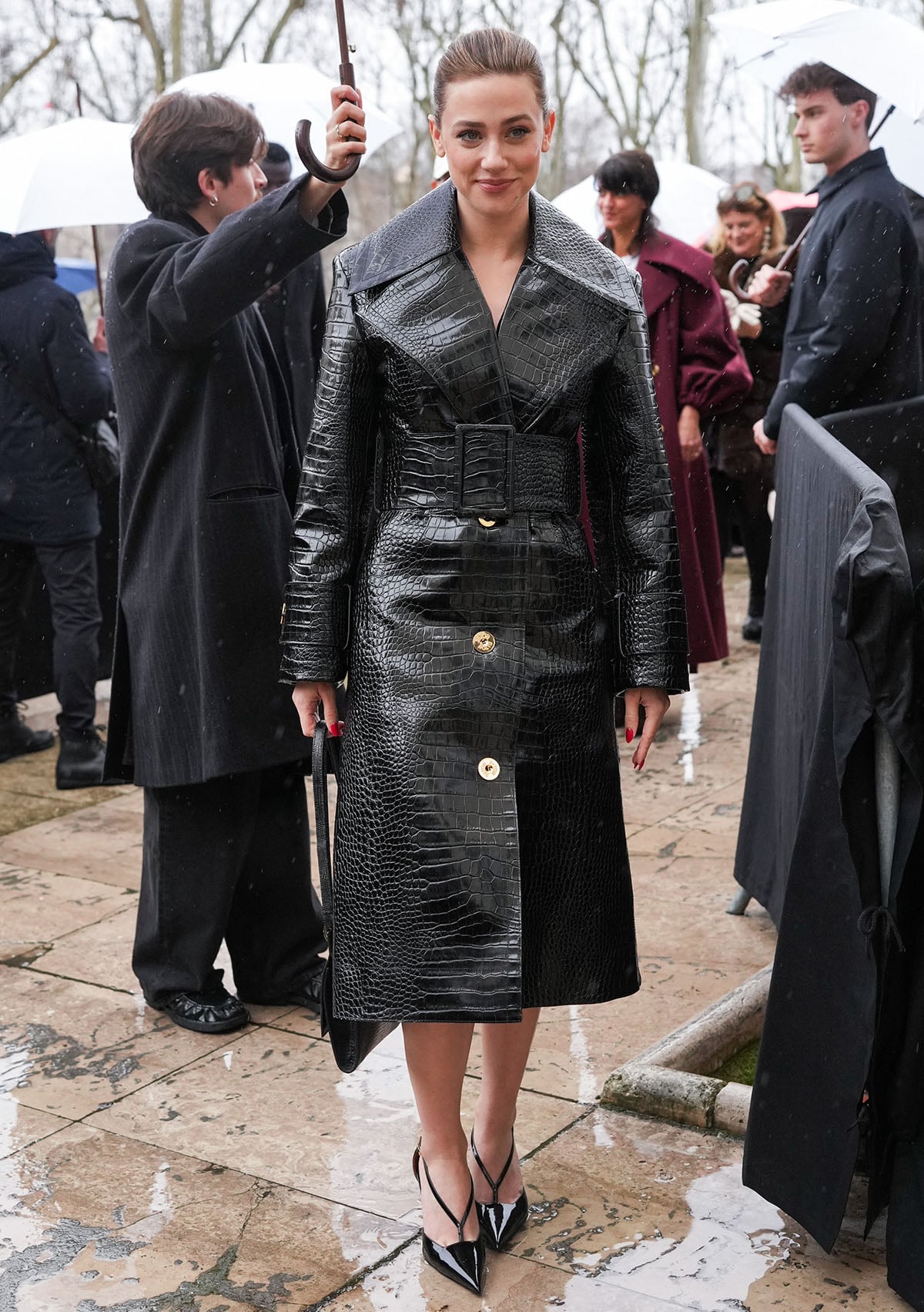 Lili Reinhart pairs her shiny shoes with an equally shiny croc-embossed leather trench coat featuring exaggerated lapels, a wide collar, gold-tone front buttons, and a wide buckled belt