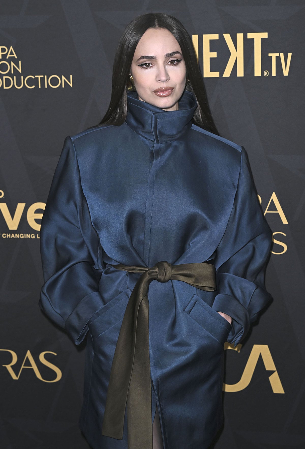 Sofia Carson opts for a modern look in an oversized blue satin Saint Laurent belted coat worn as a mini dress, featuring a high stand-up collar, sculpted bouffant sleeves, and a self-tie belt